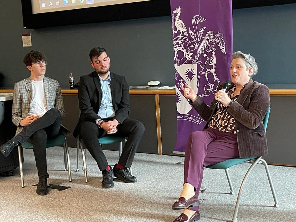 Ashley Dalton MP talking into a microphone, with two young men in smart clothing, at Edge Hill University